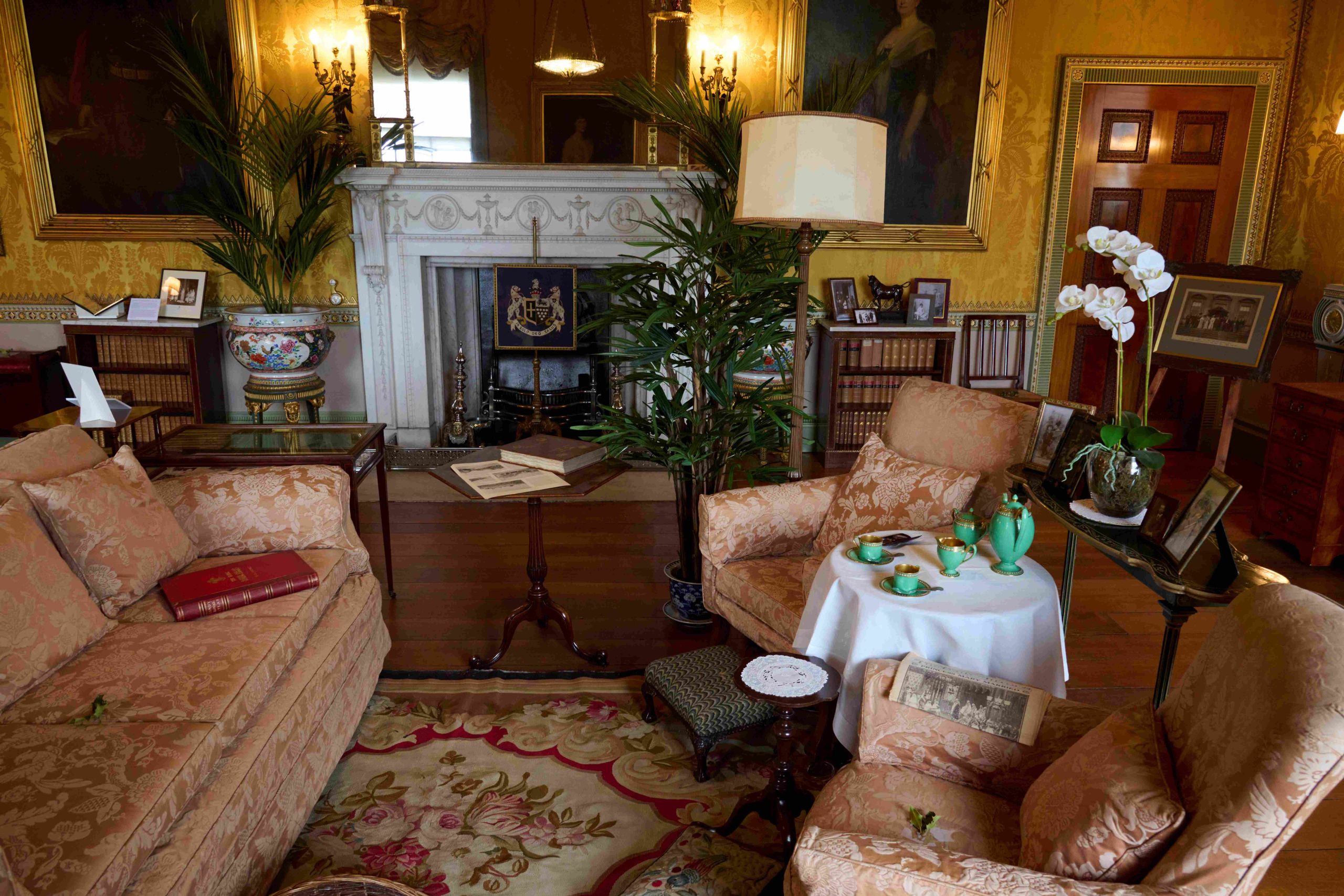 A grand interior with yellow damask walls and a large white marble fireplace. The room is filled with apricot sofas, lots of small tables and plants.