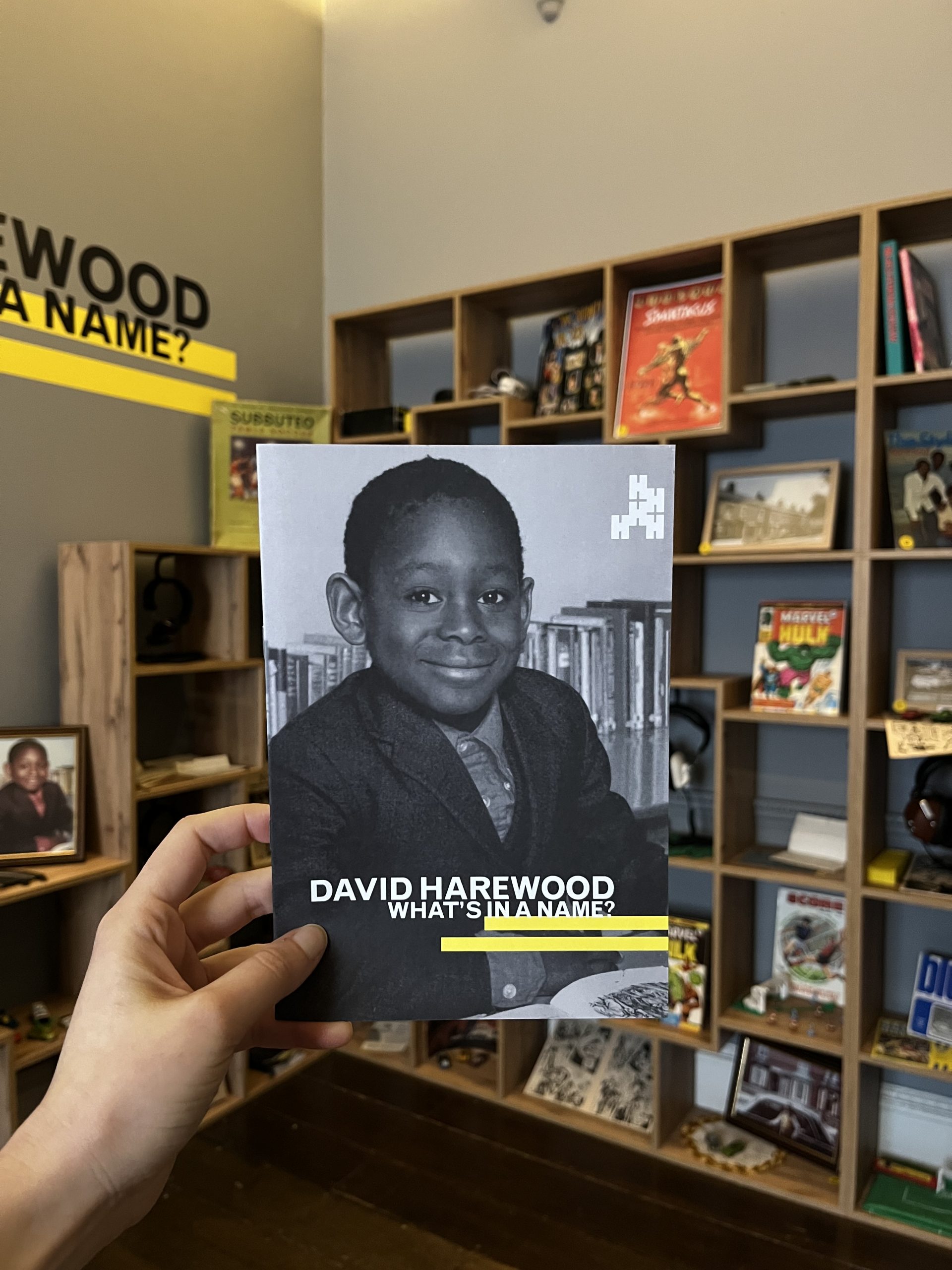 A photograph of a booklet produced for the David Harewood exhibition featuring an black and white childhood school photograph of David himself. Behind are a number of shelves containing objects representing David's childhood.