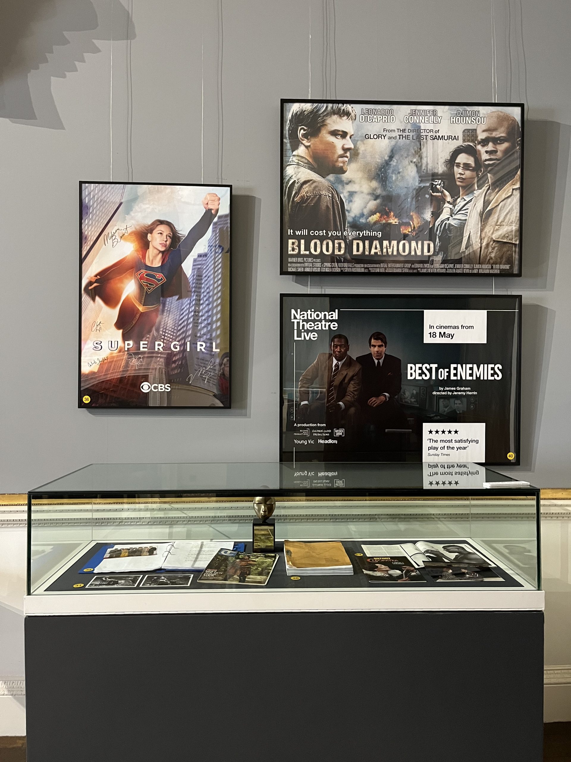 A glass-topped display case containing a number of documents and photographs from David Harewood's archive. Three large movie posters are hung above, made to advertise Supergirl, Blood Diamond and Best of Enemies.