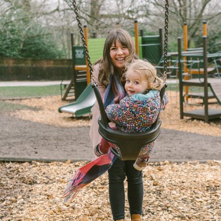 A young women with dark brown hair and a fringe smiles sweetly as she pushes her daughter on the swings in Harewood Adventure Playground.