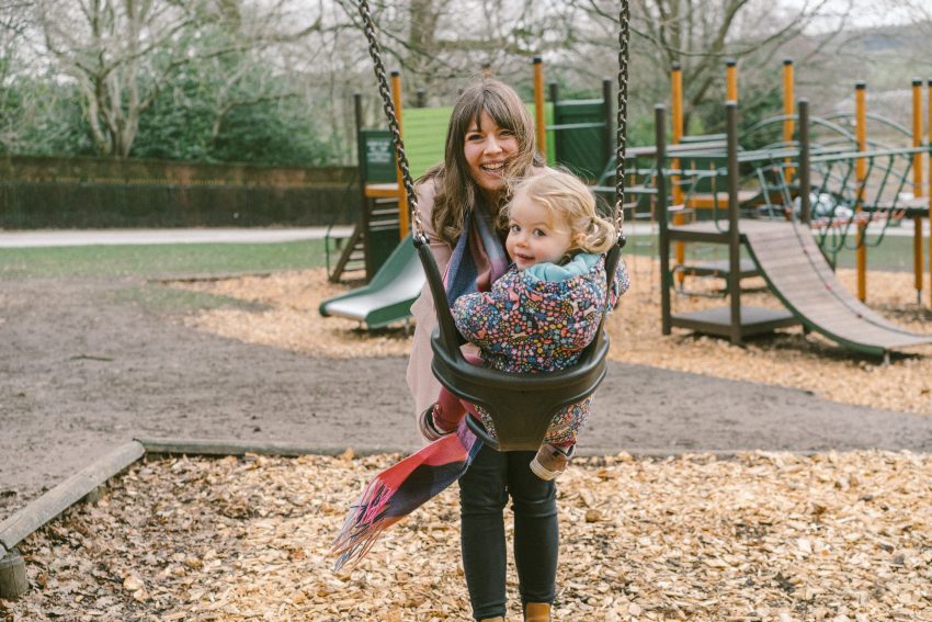 A young women with dark brown hair and a fringe smiles sweetly as she pushes her daughter on the swings in Harewood Adventure Playground.