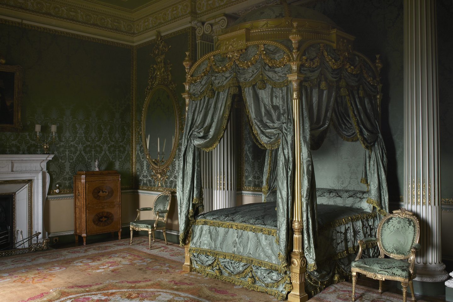 Luxurious bedroom with ornate, green and gold canopy bed, matching chairs, intricate wallpaper, rich decor, wood cabinet, and elegant rug, evoking opulence and grandeur.