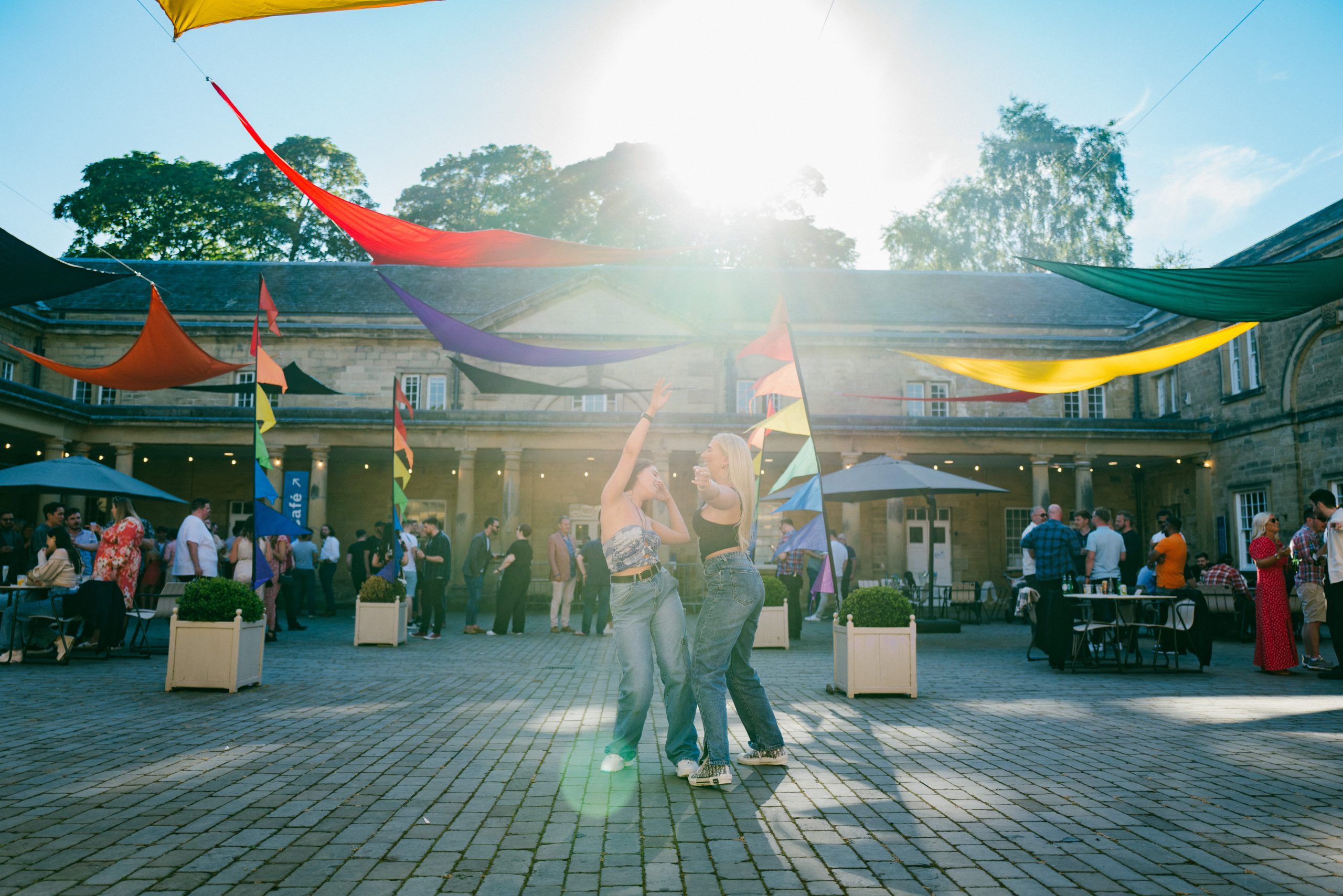 Two young women dance in the middle of the Courtyard under the late evening sun. In the background their colleagues celebrate their Summer work do at Harewood with drinks and a BBQ.
