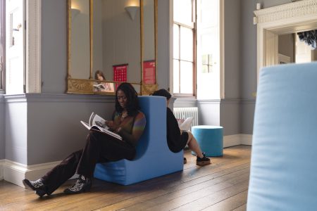 Two women read whilst sat on a blue plush chair that is shaped like an upside down T.