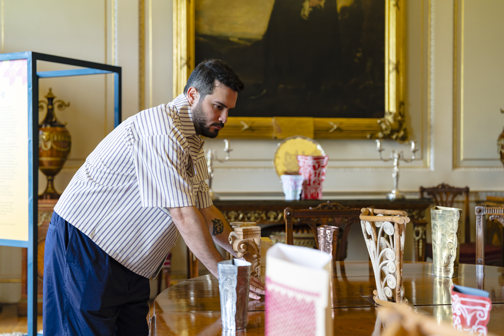 A man with dark hair and facial hair leans over a wooden table covered in corinthian style vases. The vases are made from different materials: wood, bamboo, bronze, gold, canvas and leather.