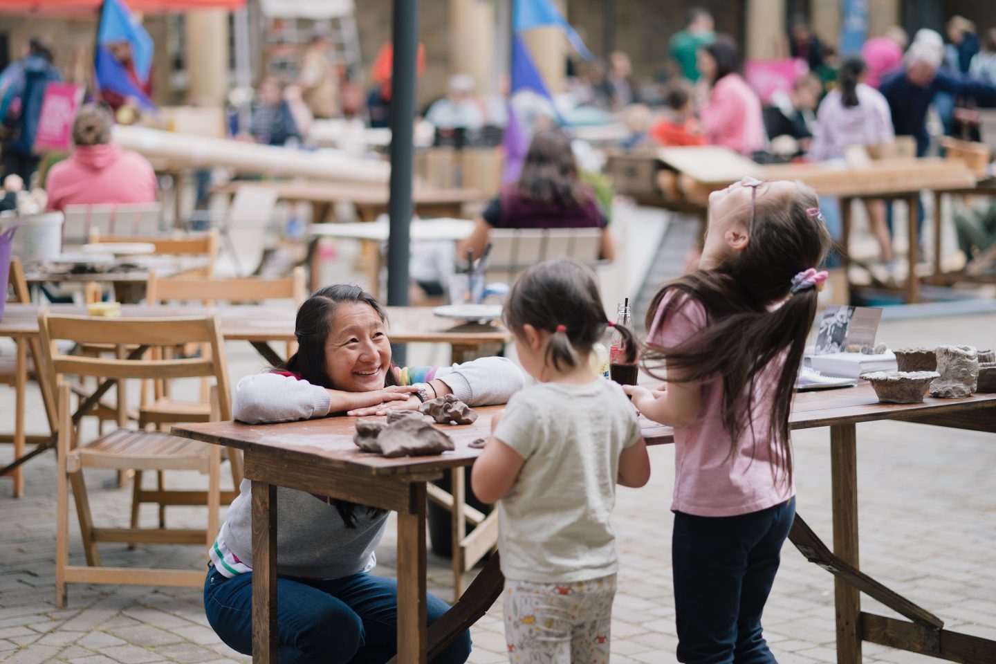 A women crouches at a wooden table smiling as she watches her two children laugh and play with modelling clay in Harewood Courtyard.