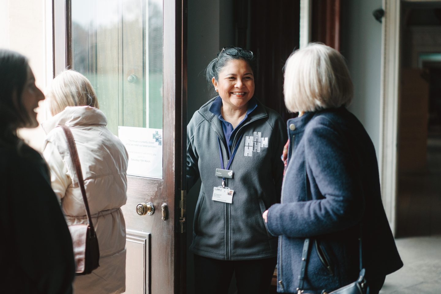 A happy women with dark hair and glasses balanced on her head holds the door open for visitors as they leave the House.