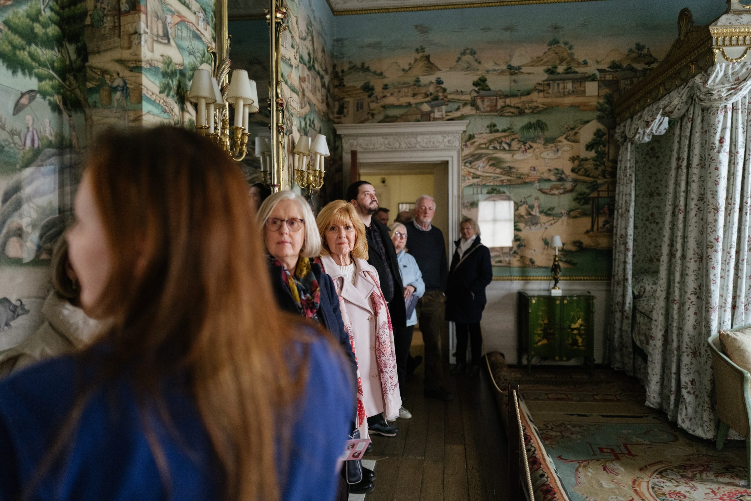A group of people line the walls of the East Bedroom listening to a tour delivered by a young member of staff.