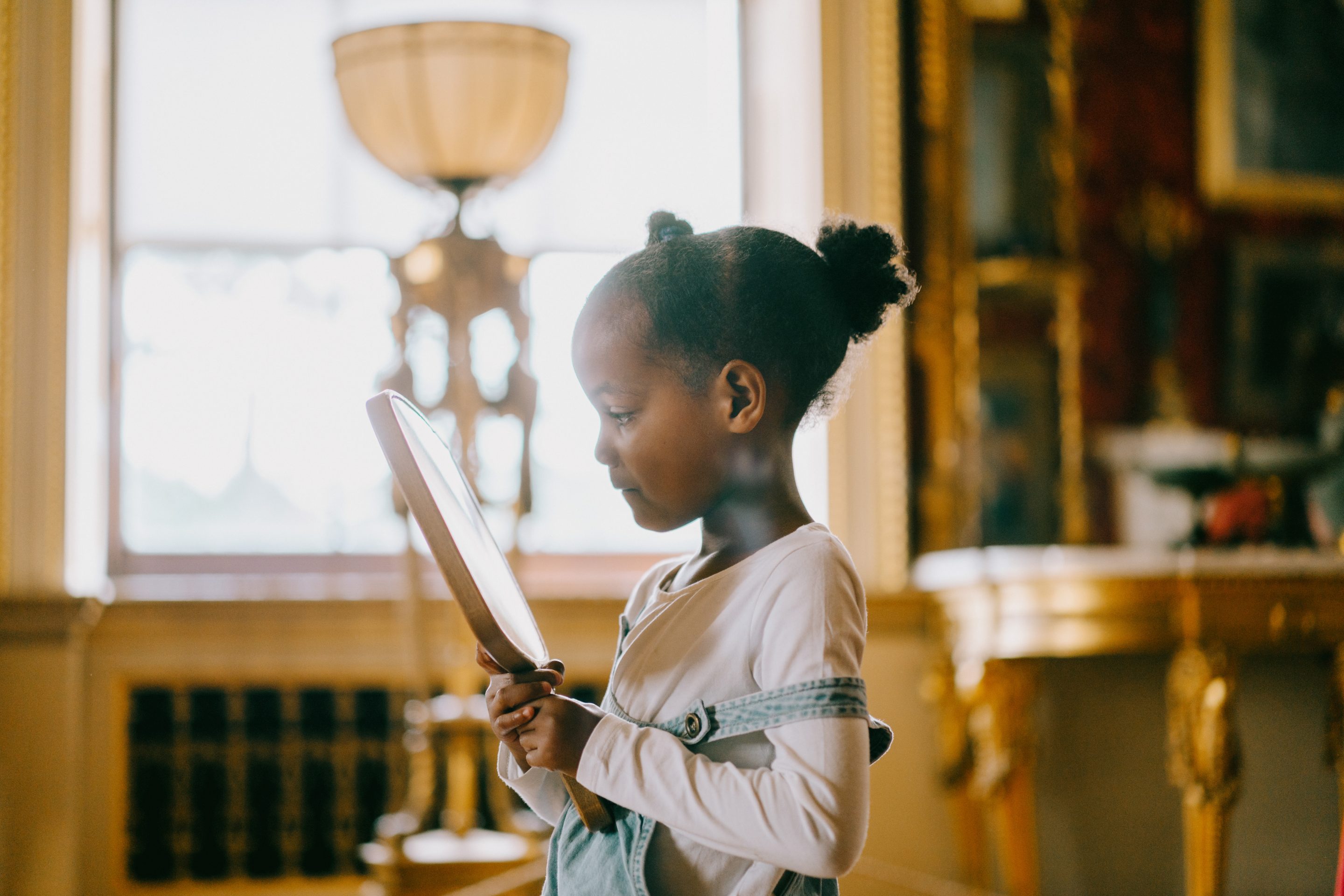 A young girl with dark brown hair pulled into a bun, looks into a hand held mirror. The girls stands in the middle of the long Gallery near a window.