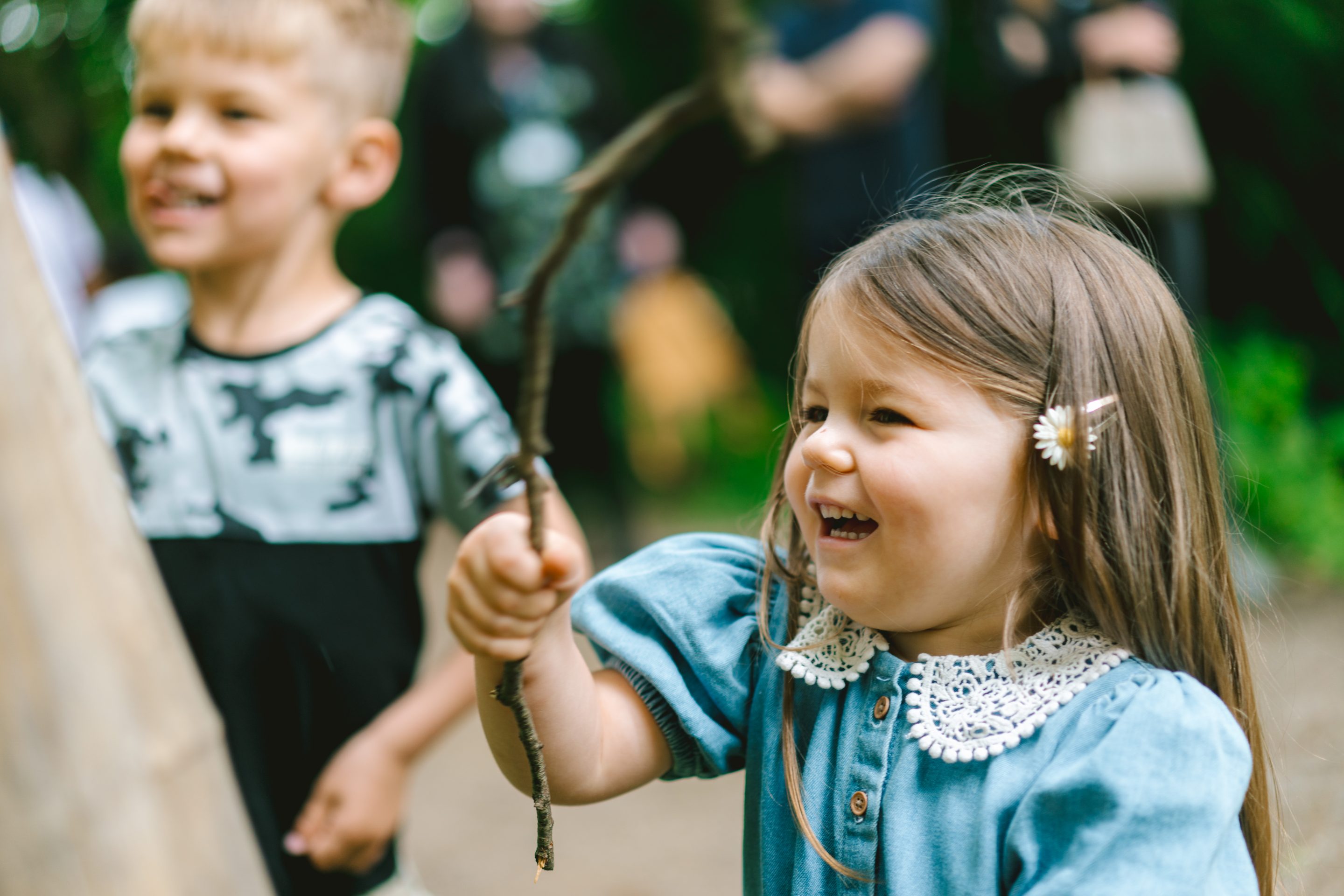 A young girl wearing a denim blue playsuit with a white crochet collar, waves a stick she found along the Lakeside path.