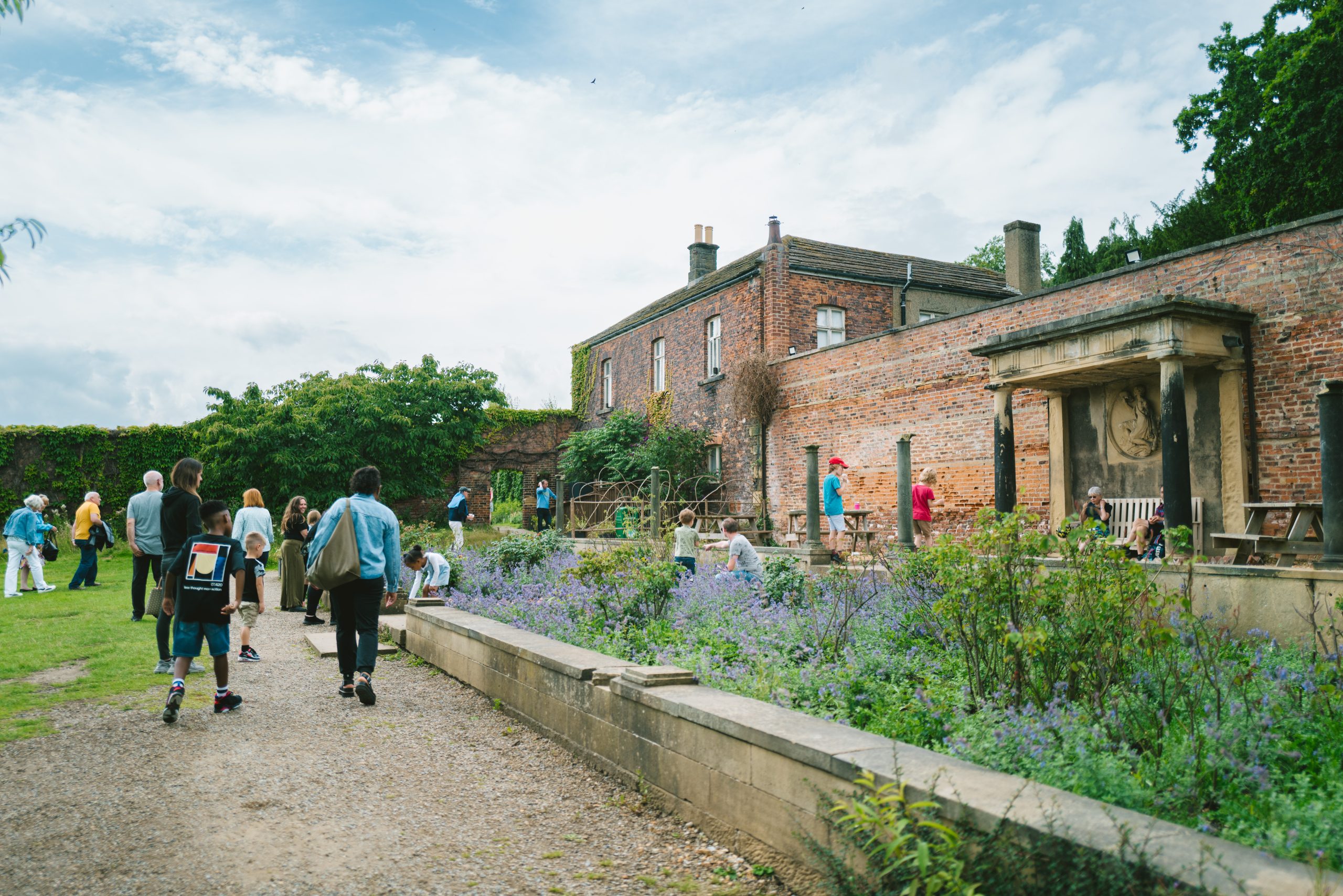 A group of Harewood visitors walk alongside the lavender garden towards the Bothy Teahouse. Ivy grows alongside walls of the Walled Garden in the background.