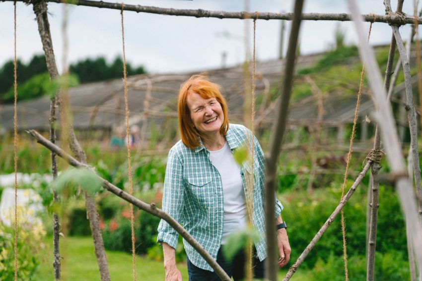 A women with ginger shoulder length hair and a fringe smiles as she looks through a structure of garden twine and willow branches with beans growing below.