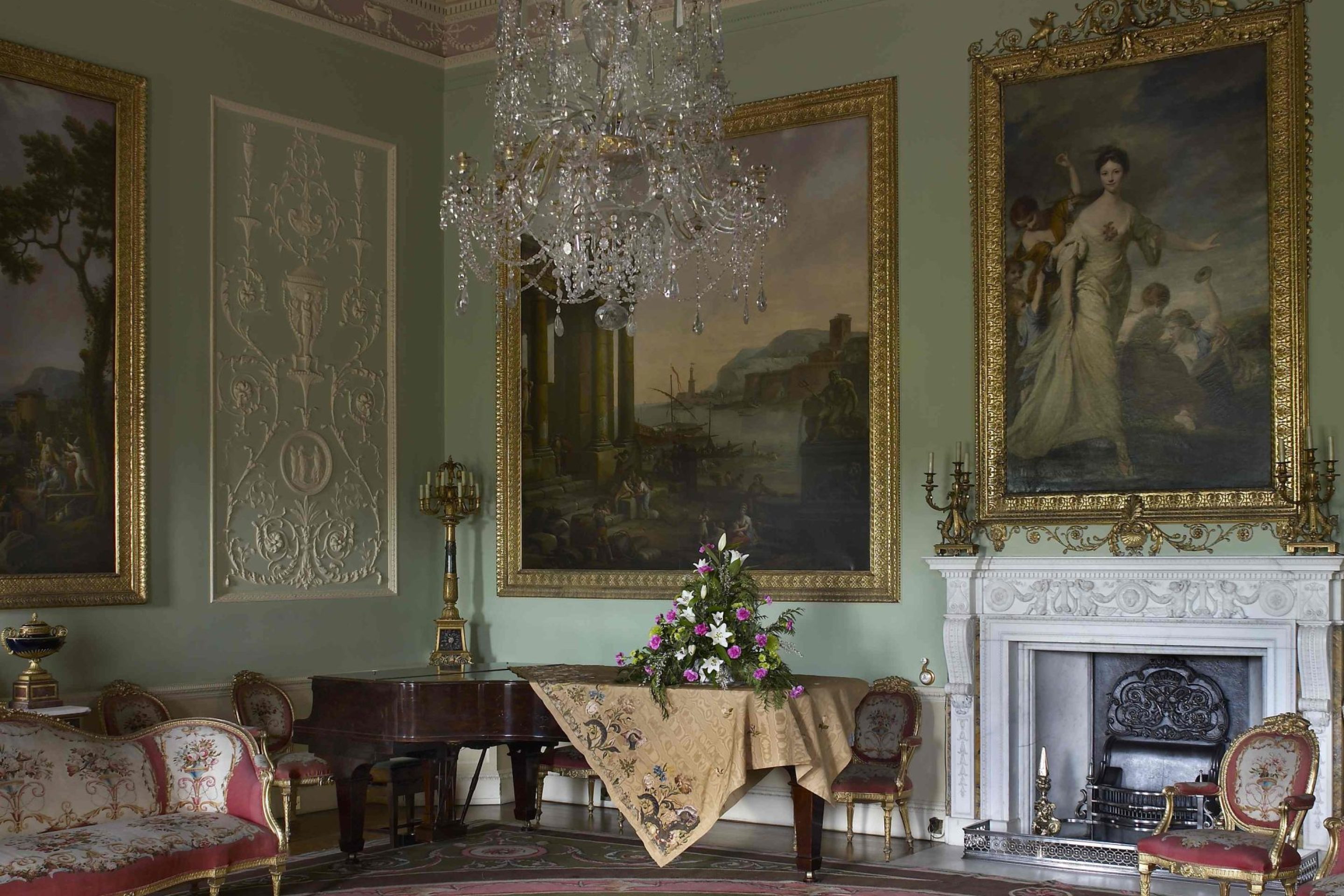 A large 18th century room with delicate plasterwork that looks like icing around the room. The walls are decorated with large oil paintings and a pink and green carpet with similar patterns to the walls covers the floor. There is a grand piano in the corner of the room.