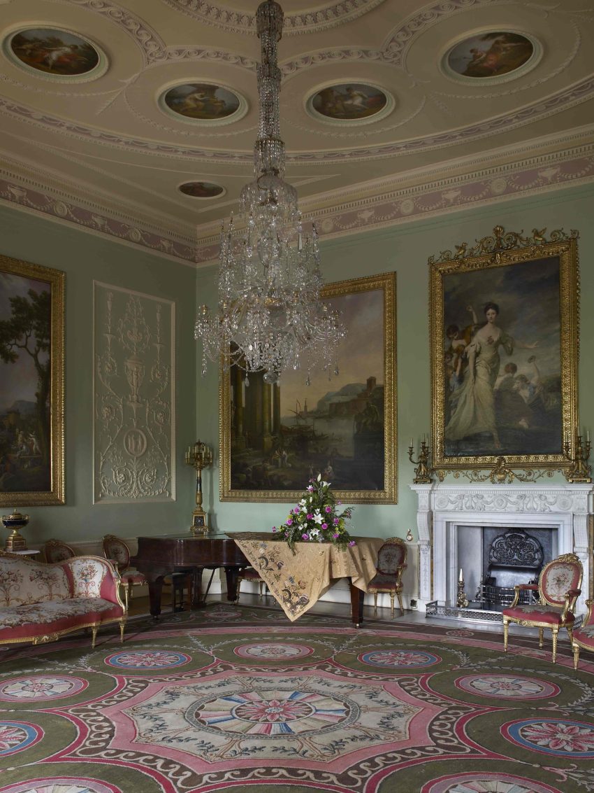 A large 18th century room with delicate plasterwork that looks like icing around the room. The walls are decorated with large oil paintings and a pink and green carpet with similar patterns to the walls covers the floor. There is a grand piano in the corner of the room.