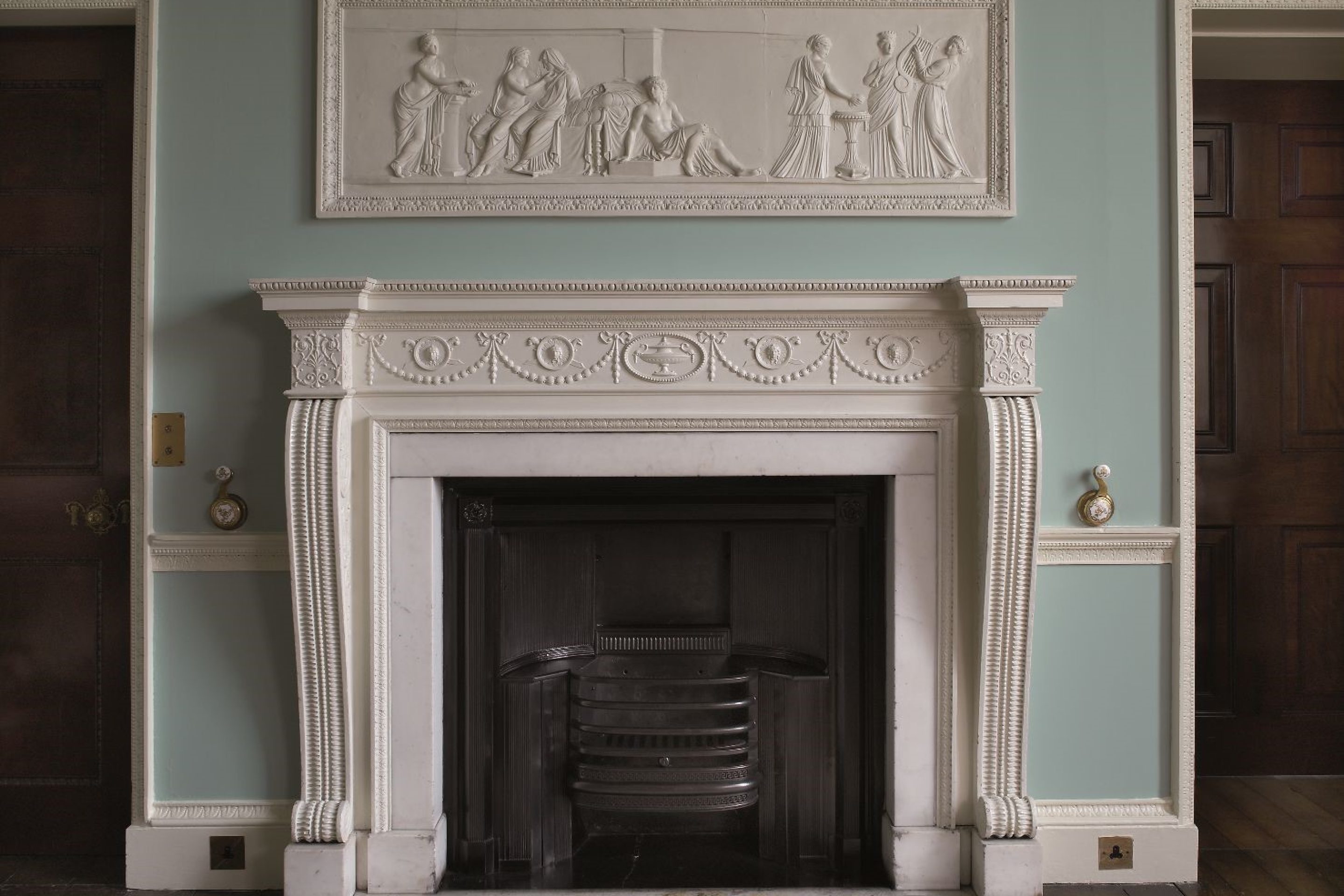 A white, intricately carved mantlepiece below a plasterwork frieze depicting a classical scene.