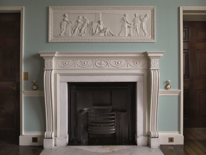 A white, intricately carved mantlepiece below a plasterwork frieze depicting a classical scene.