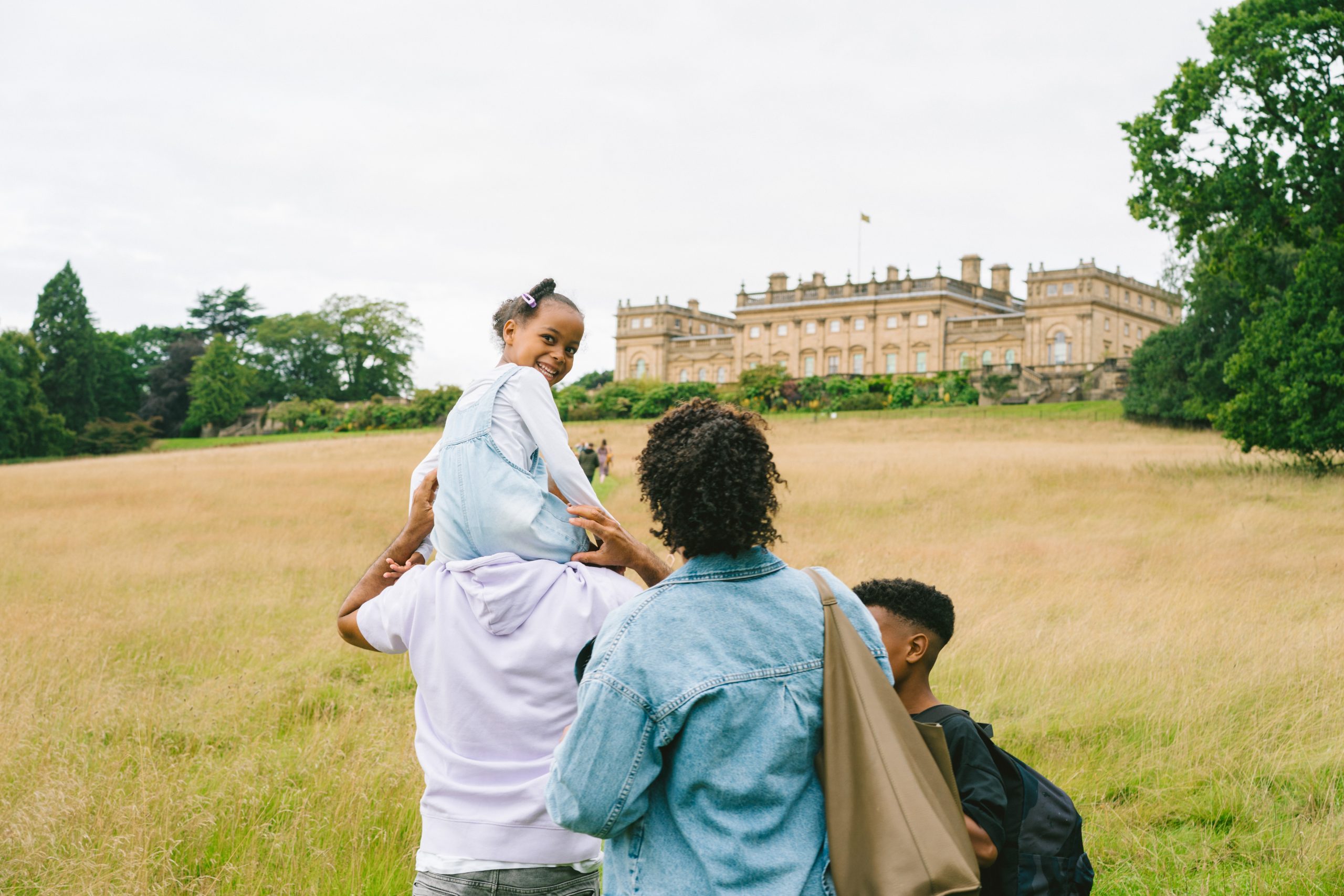 A young family walking the south park walk up towards the Archery Border and Terrace. A young girl, carried on the shoulders of her father, looks over her own shoulder to smile at the camera
