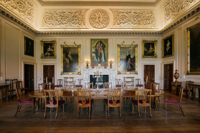 A large room with cream walls and dark wooden floorboards. In the middle of the room stands a large mahogany dining room table surrounded by 12 wooden chairs with deep leather cushions. Large gilded frames cover the walls with paintings of those who once lived at Harewood.