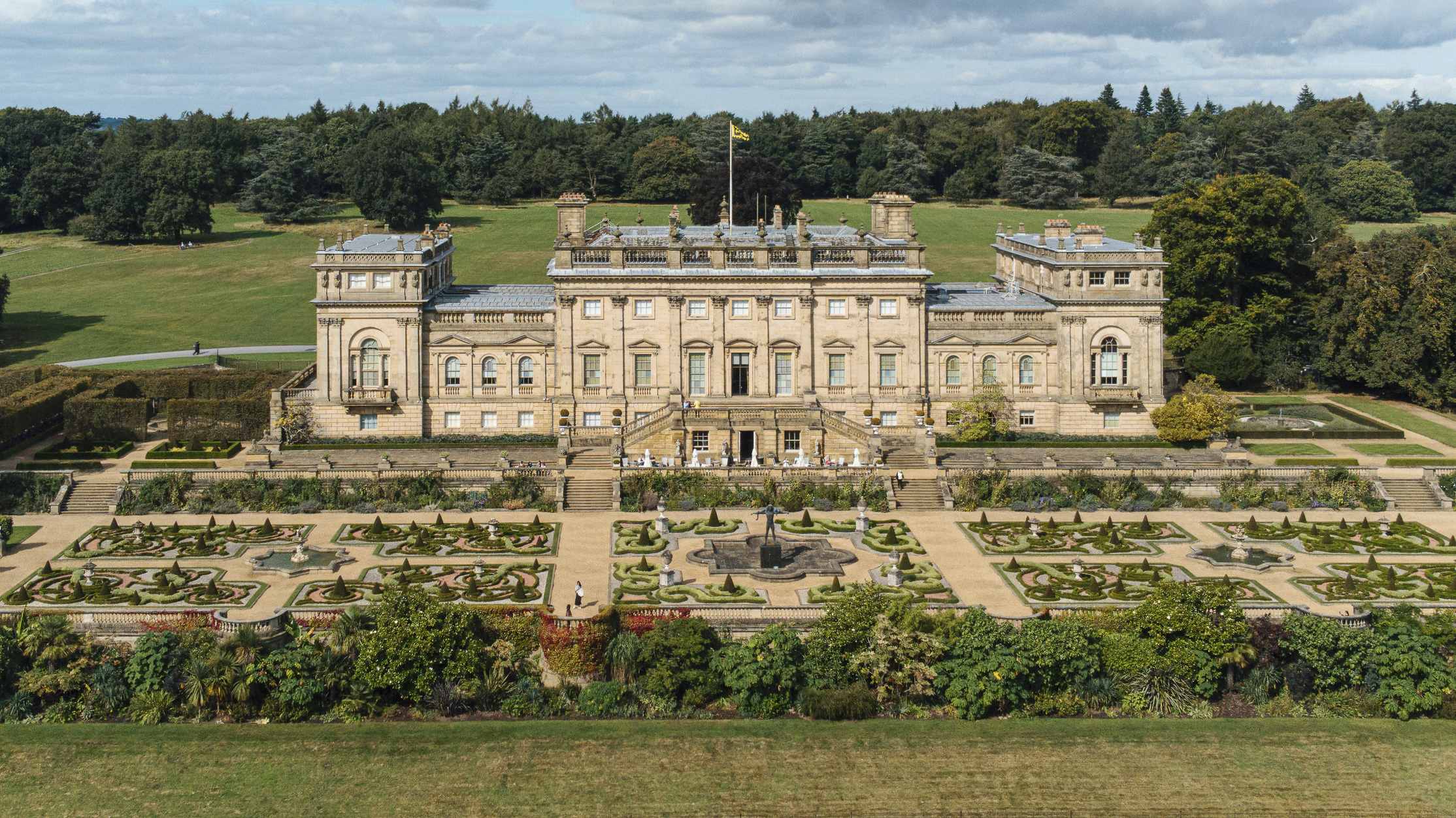 Aerial view of Harewood House, a grand, symmetrical estate building with classical architecture, featuring tall columns and large windows, set against a backdrop of lush, green lawns and dense trees. In the foreground, an italianate parterre displays intricate patterns of hedges and paths surrounding a central fountain.