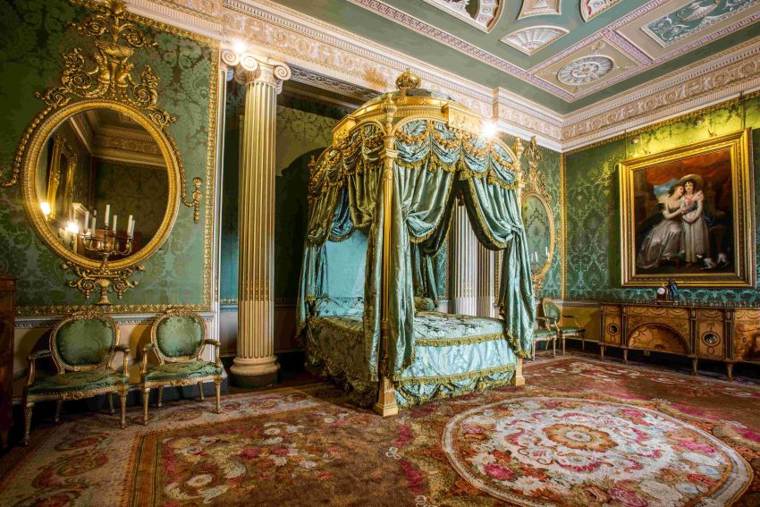 A bedroom upholstered in rich green damask and decorated with a pair of gilded mirrors. A large gilded four-poster bed stands in the centre.