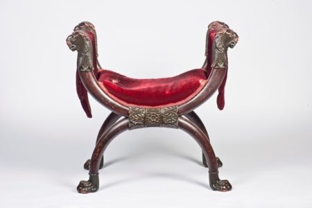 A painted stool with an x-shaped frame and a red velvet cushion. The legs are shamed like large lion paws with the head of lions on the arm rest.