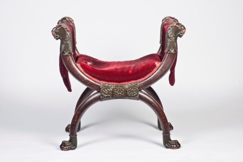 A painted stool with an x-shaped frame and a red velvet cushion. The legs are shamed like large lion paws with the head of lions on the arm rest.