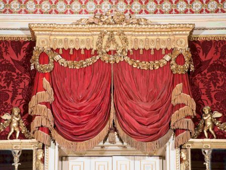 A large gilded pelmet with red drapes carved out of wood.