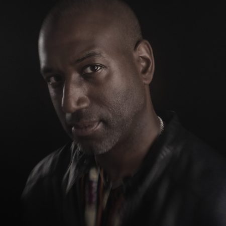 A man wearing a black jacket tilts his head to the camera. The light reflects in his eyes and bounces from his silver necklace.