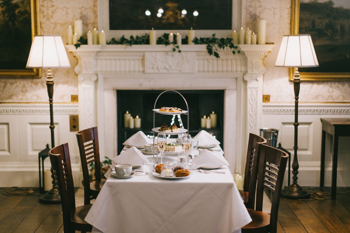 Four wooden chairs surround a table dressed for Afternoon Tea, with glasses of Nyetimber sparkling wine, scones, sandwiches and sweet treats. The table stands in front of an opulent fireplace decorated pillar candles and ivy.