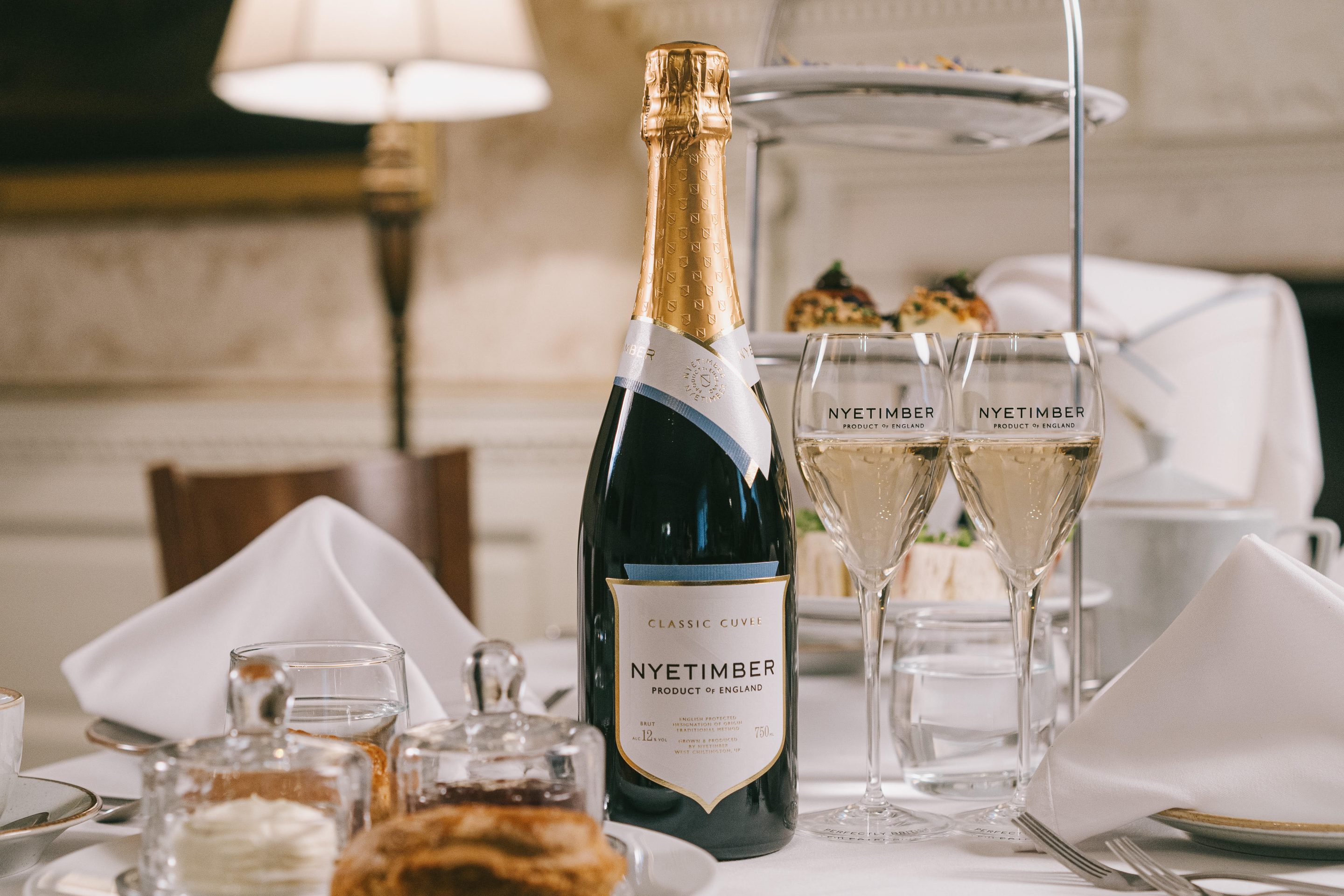 Table with white table cloth laid up for Afternoon Tea with Nyetimber sparkling wine. Scone, cream and jam sits in the foreground with three tiers of plates, each tier containing a different type of delicious food stands in the background.