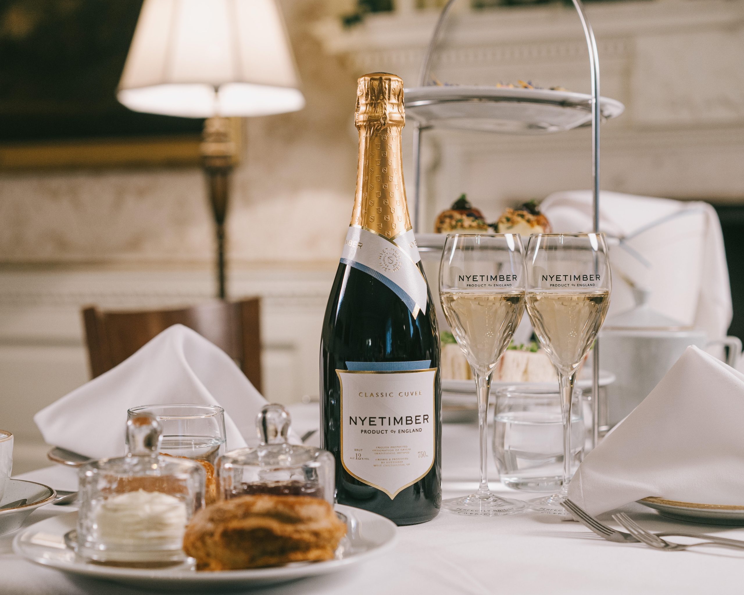 Table with white table cloth laid up for Afternoon Tea with Nyetimber sparkling wine. Scone, cream and jam sits in the foreground with three tiers of plates, each tier containing a different type of delicious food stands in the background.