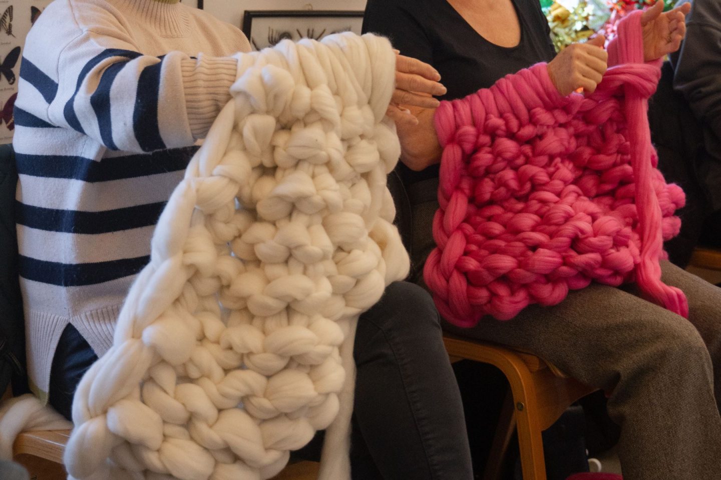 Two ladies have yarn wrapped around their arms. The lady on the left is using white wool and the lady on the right is using bright pink. They are using their arms like knitting needles and a blanket has formed underneath their arms.