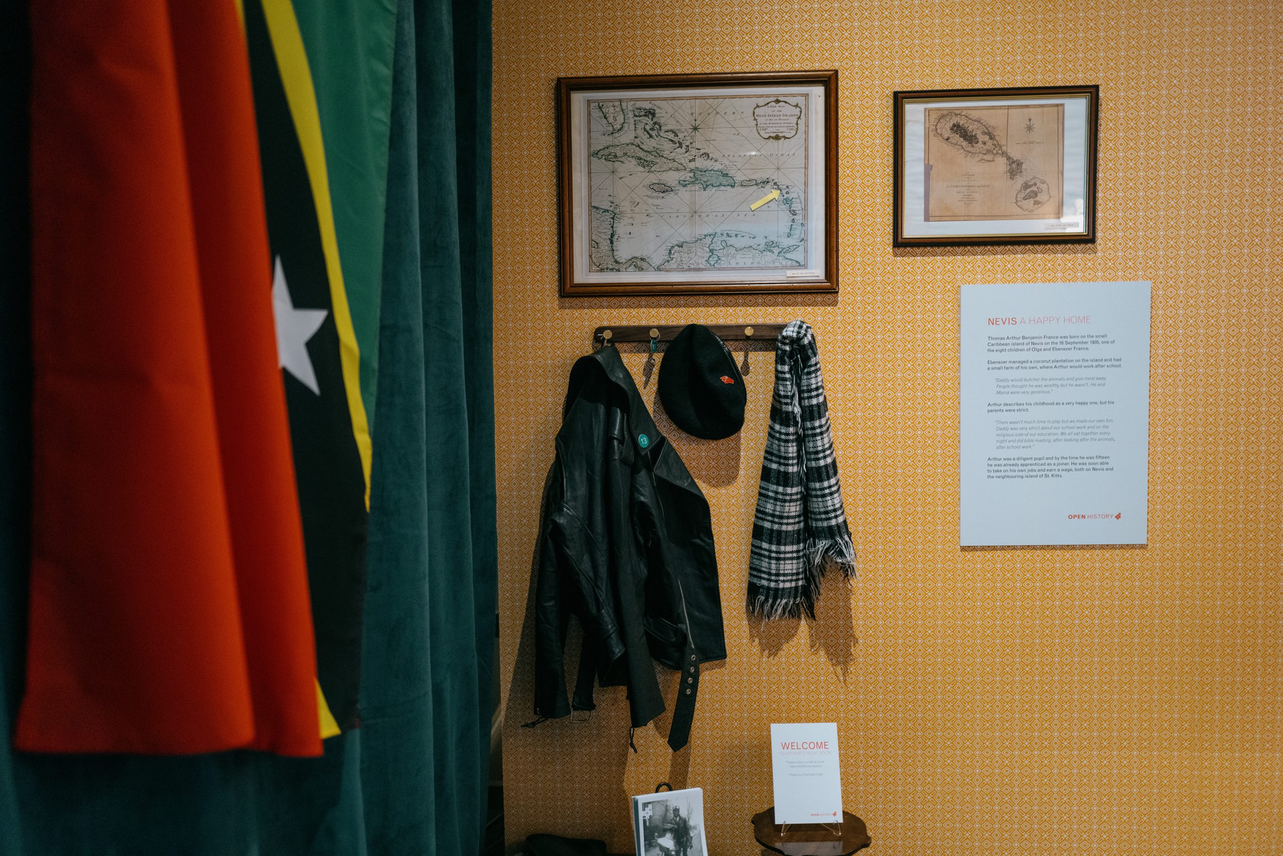 A map of Caribbean Islands with a small yellow arrow pointing to the island of Nevis hangs on a wall above a coat hook were a black beret, monochrome check scarf and black leather jacket hangs. The flag of Saint Kitts and Nevis hangs on the adjacent wall.