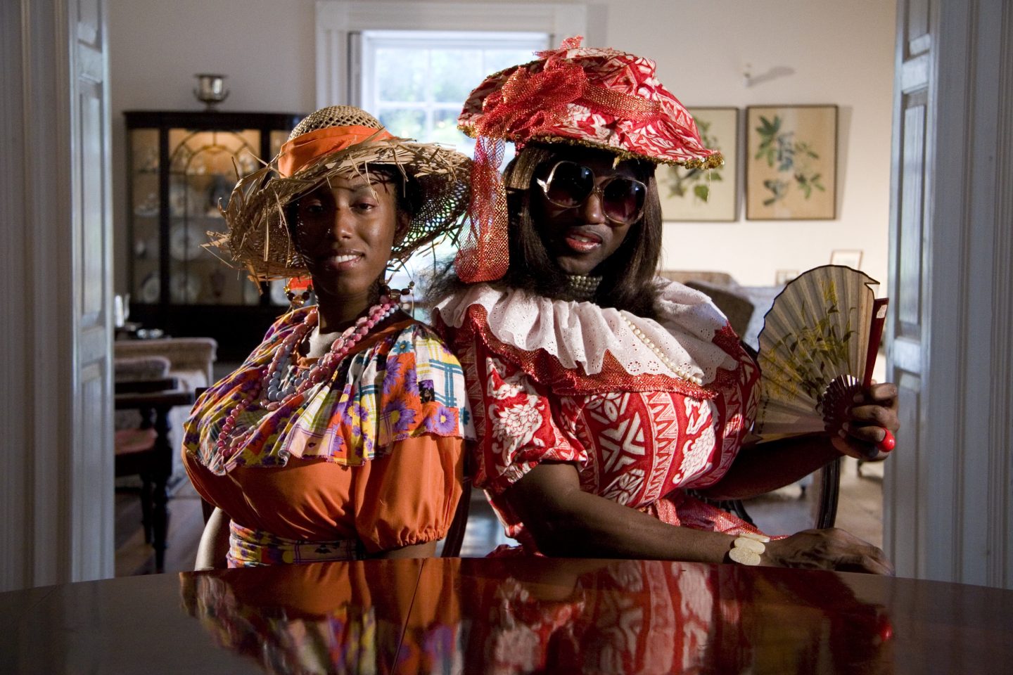 Two people wearing bright, patterned dresses, one in red, one in orange and matching straw hats. One people wears big sunglasses and holds a fan, the other wears thick beaded necklaces. They are sat at a mahogany table smiling directly at the camera.