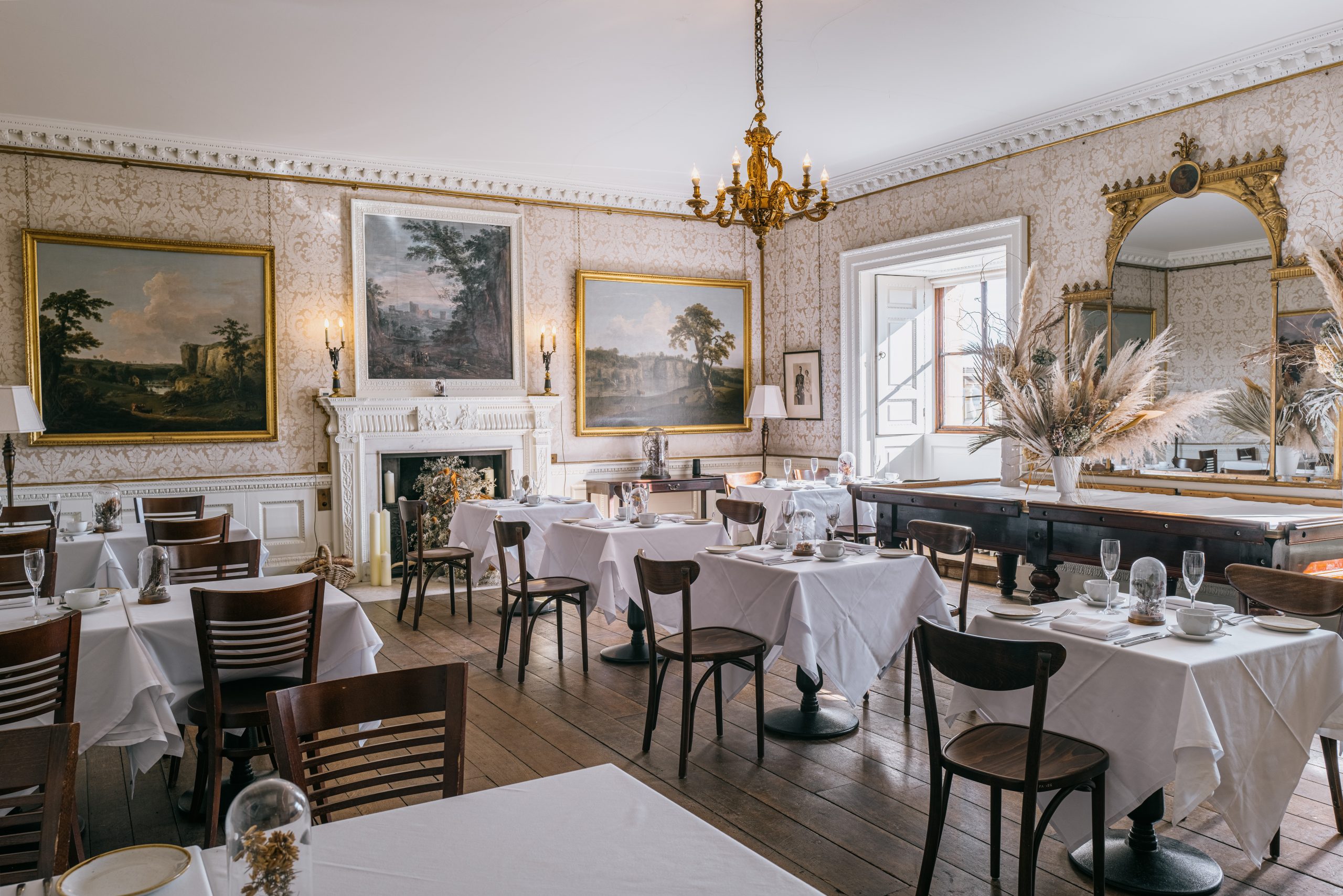 Elegant dining room with white tablecloths, wooden chairs, and a chandelier. Walls are adorned with Harewood landscape paintings by J. M. W. Turner, and a large gilded mirror reflects the room's decor.