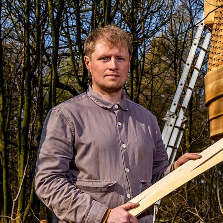 A man in a grey jacket stands in a wooded area, holding wooden planks. A partially built wooden tree house and a ladder are visible in the background.