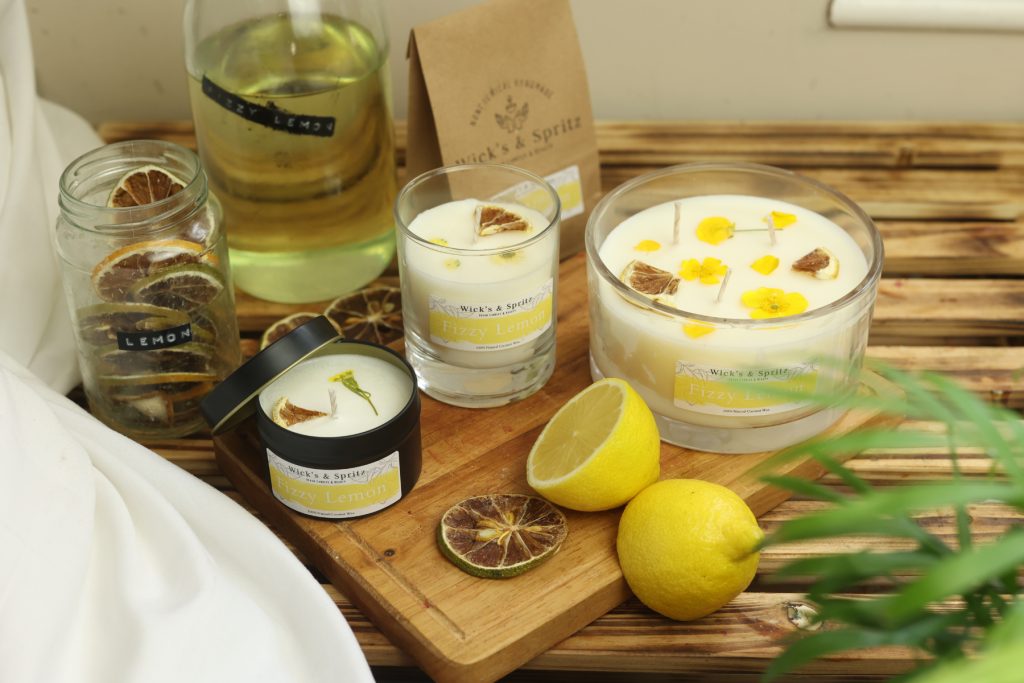 three candles laid out on a wooden chopping board. All the candles are decorated with yellow petals and dried slices of lemon. In the background there is dried lemon slices in a jar and also a yellow liquid in bottle labelled Fizzy Lemon.