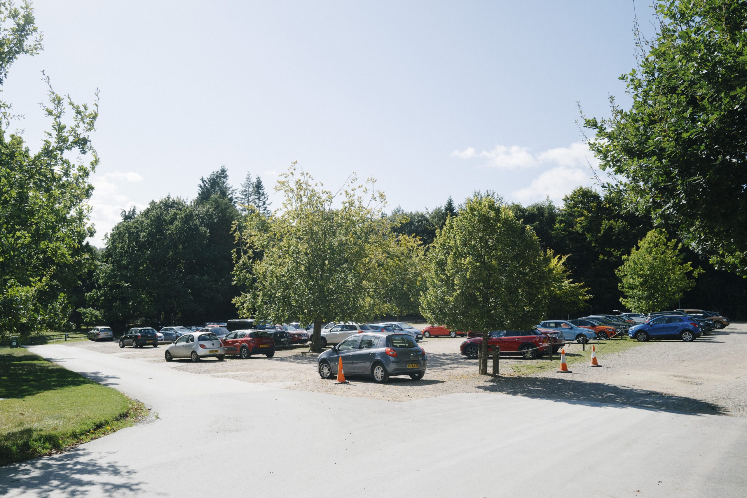 Open gravel car park, with cars parked sporadically in bays across 8 rows. Trees and greenery surround the car park.