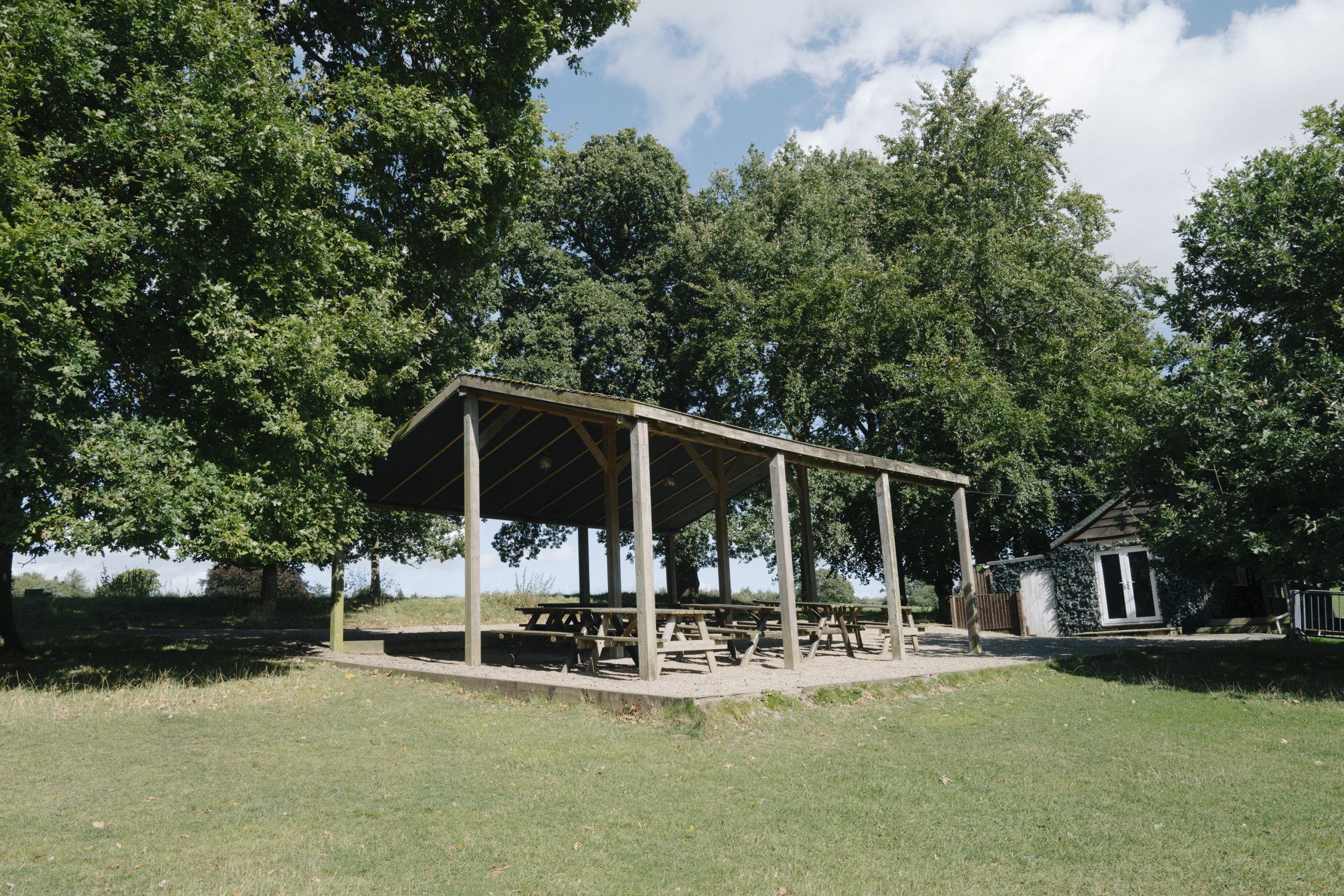 Wooden picnic benches stand on a graveled area beneath a large wooden pergola. The picnic area is surrounded by tall lush trees. A small cabin stands to the right hand side.