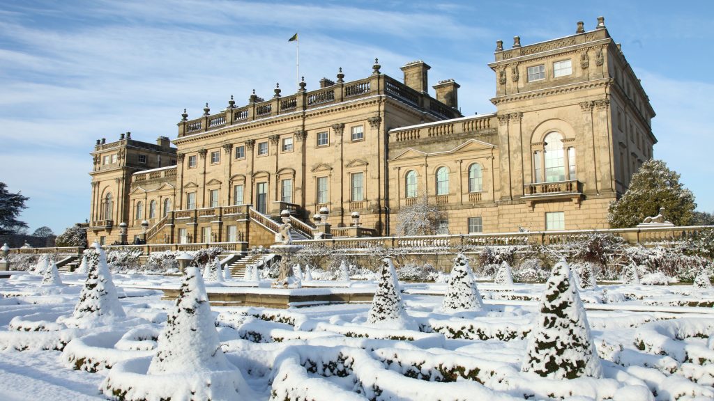 A grand, historic stone country house in neoclassical style, surrounded by formal gardens covered in a blanket of snow.