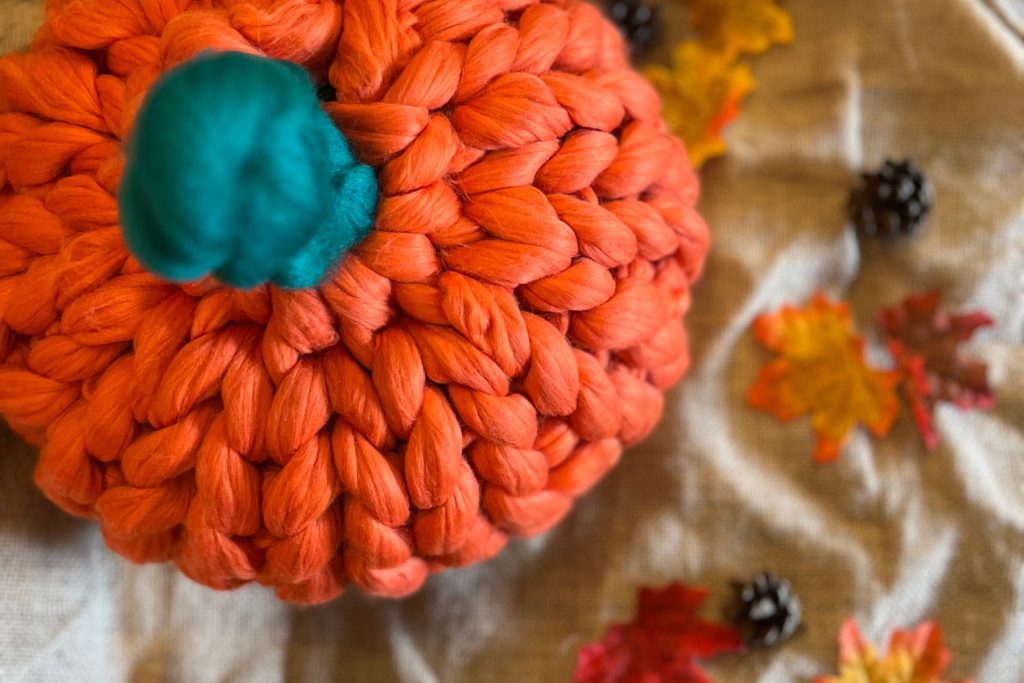 A pumpkin cushion created with bright orange wool. The pumpkin is sitting on a pile of autumn leaves and looks super squishy and cosy.