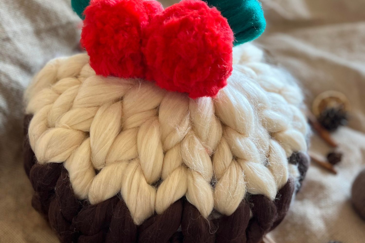 A Christmas pudding cushion made from chunky wool. The top of the christmas pudding is made from white wool to represent the icing and the rest is a brown colour. It has two fluffy red pompoms as berries and a green bow on the top.