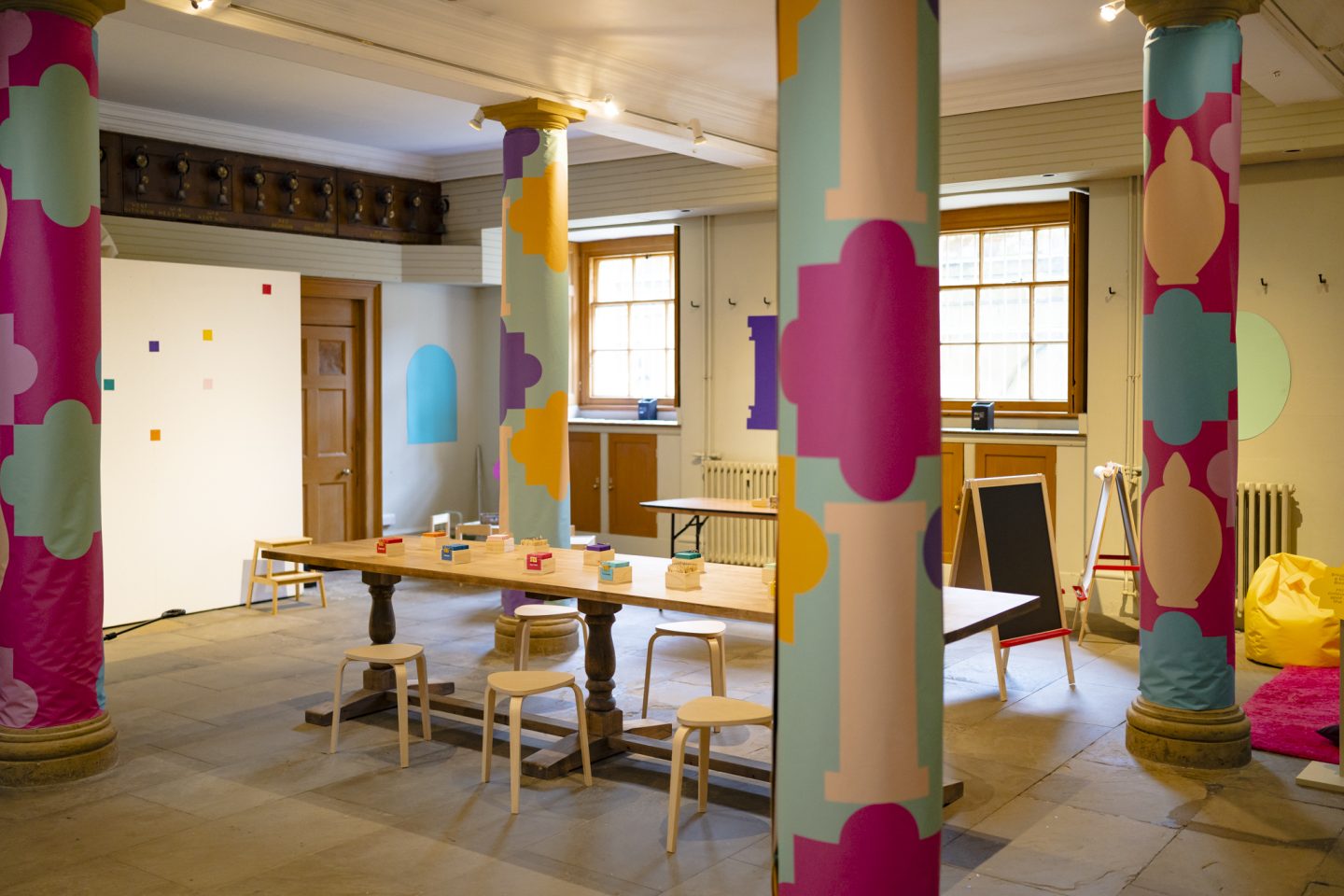 Four pillars are covered in colourful shapes. In the middle of the room stands a long wooden table surrounded by small wooden chairs and bright colourful boxes laid on top.