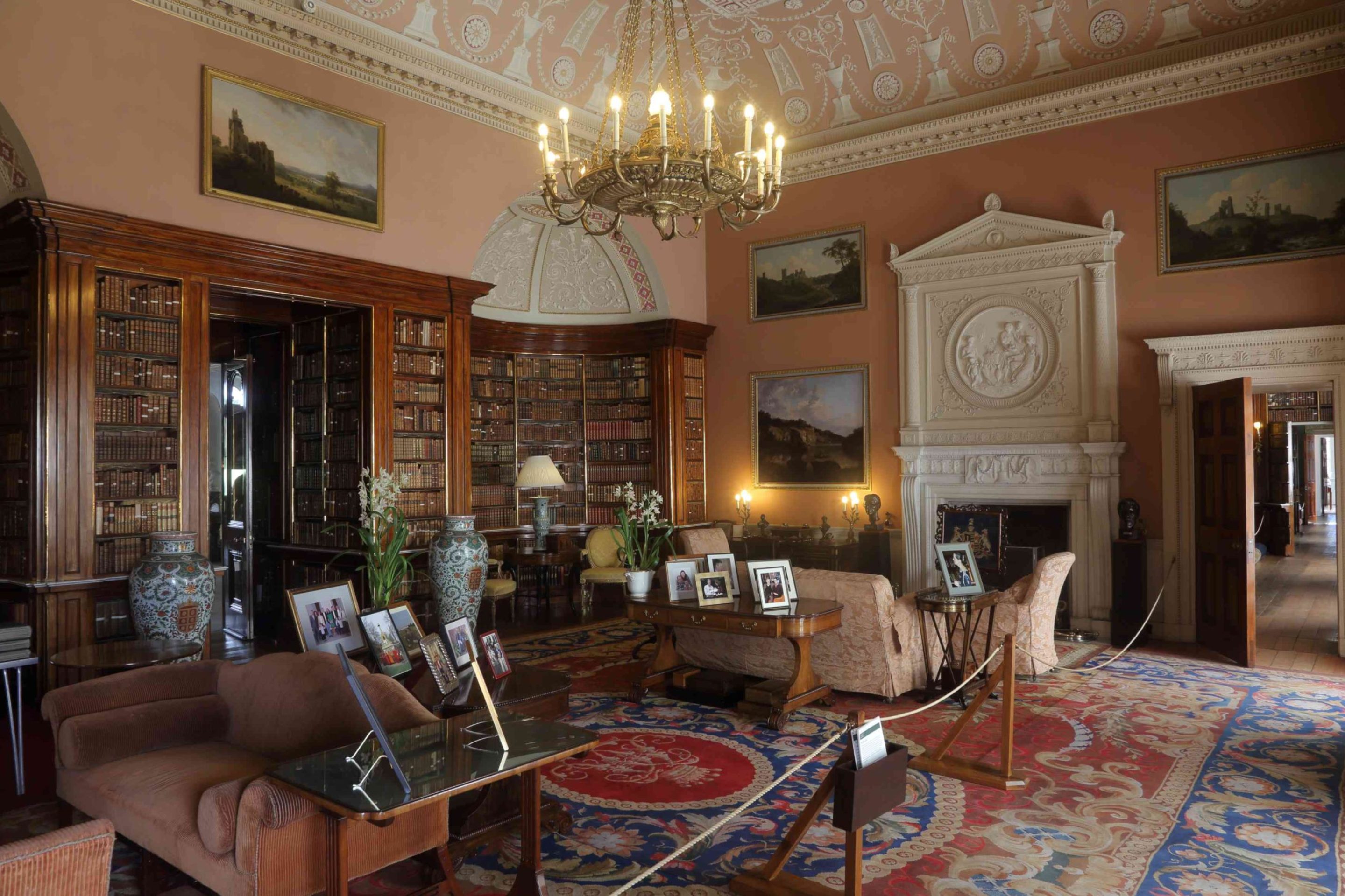 A large room with intricately decorated plasterwork ceiling and a fireplace with a pediment-shaped overmantel. On the left is an alcove entirely filled with mahogany bookcases. The room is filled with furniture and family photographs.