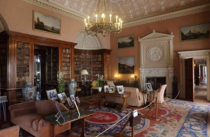 A large room with intricately decorated plasterwork ceiling and a fireplace with a pediment-shaped overmantel. On the left is an alcove entirely filled with mahogany bookcases. The room is filled with furniture and family photographs.