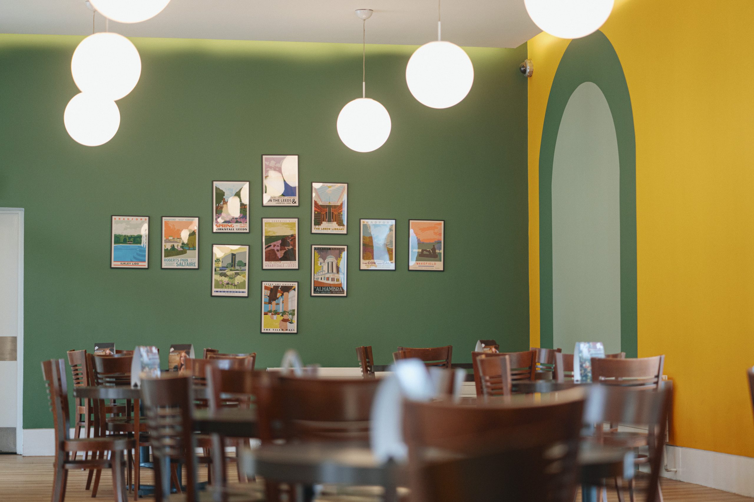 A modern cafe seating area with dark wooden chairs and tables, featuring a forest-green wall covered in colourful framed posters of various Yorkshire landscapes and landmarks. A mustard-yellow accent wall with a curved arch complements the decor, while circular pendant lights provide soft lighting.