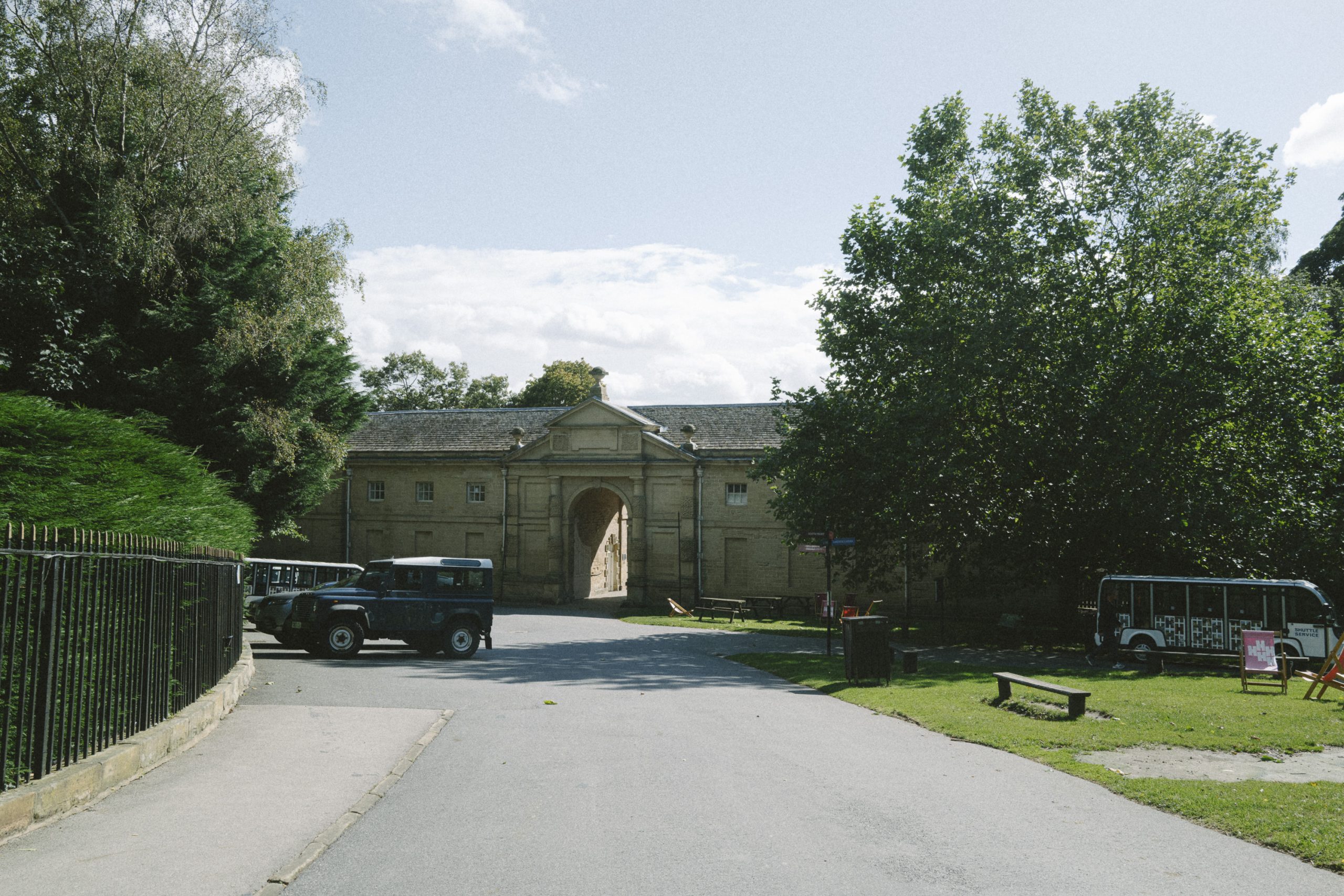 A short paved road leads down to the old stable yard, known as the Courtyard. Disabled parking spaces are filled with cars just left of the Courtyard, with Harewood Shuttle Bus parked at the bus stop to the right. Lush large trees grow either side of the road with bright blue skies overhead.