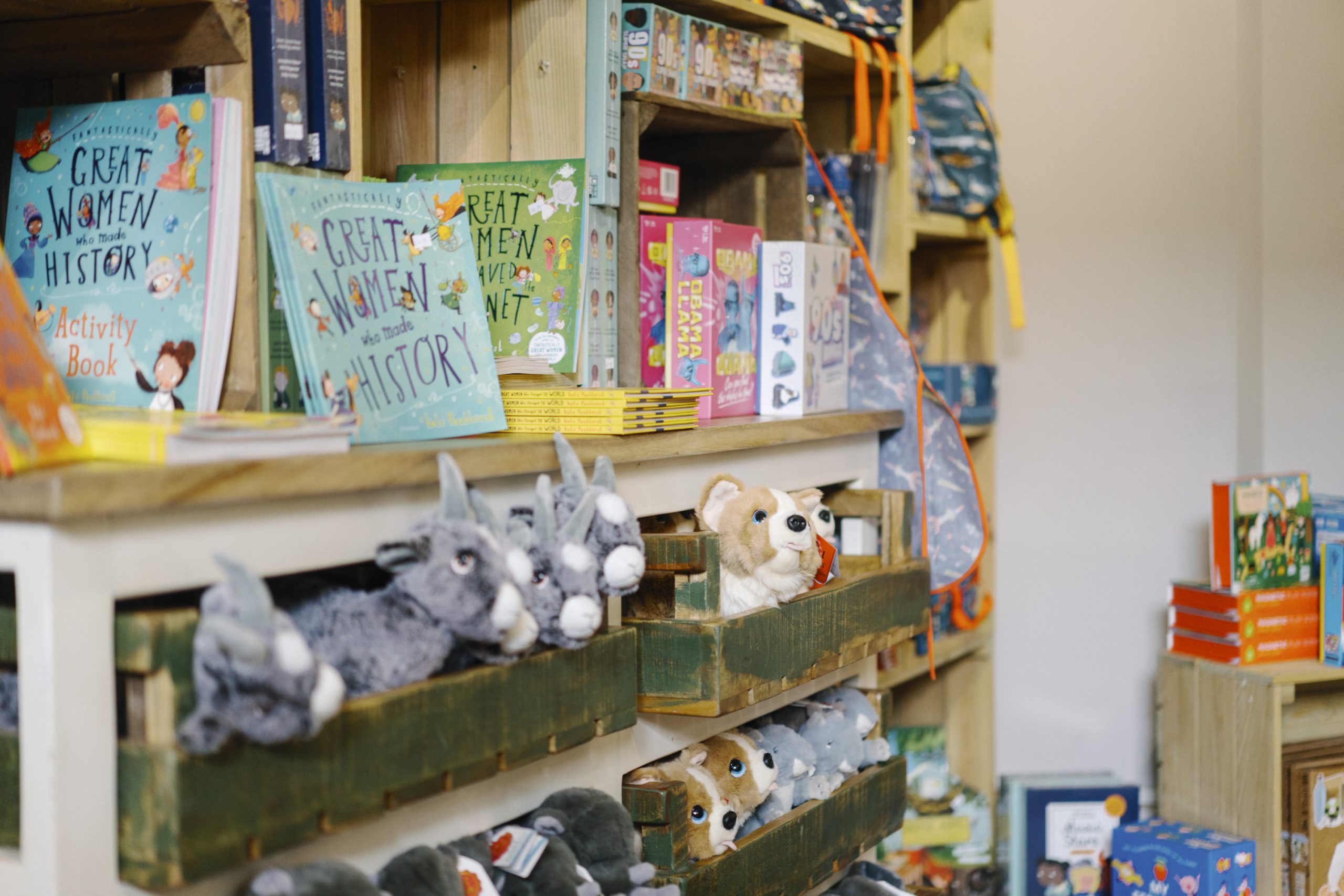 A coluorful retail display in the Courtyard Shop featuring children's books and plush toys. Books titled Great Women Who Made History are prominently displayed on wooden shelves, alongside stuffed animal toys in rustic wooden bins. Backpacks and additional merchandise are arranged in the background.