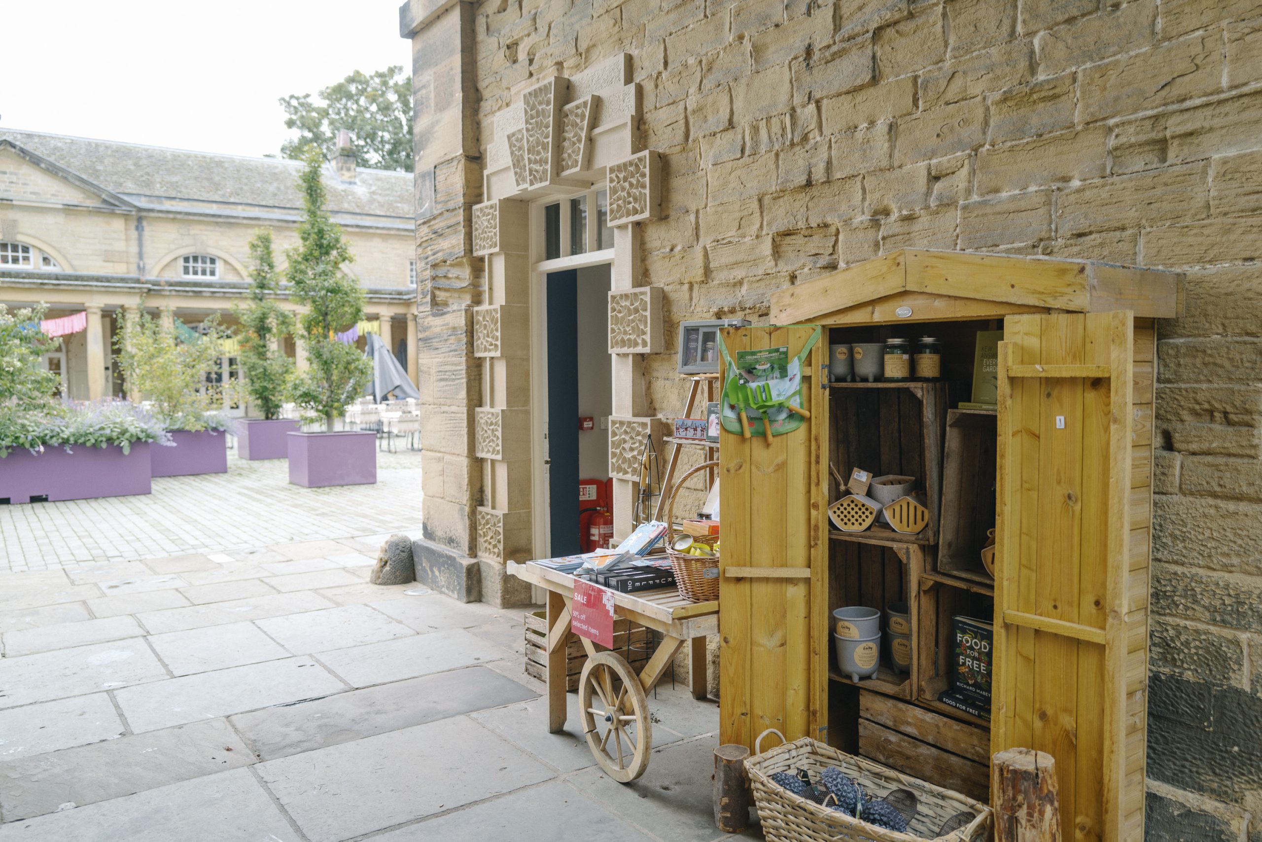 A small wooden shed outside the Courtyard Shop, stocked with gardening gifts such as acorn-shaped bird feeders, plant pots, and insect hotels. In the softly blurred background, the Courtyard seating area features new trees planted in large purple planters.