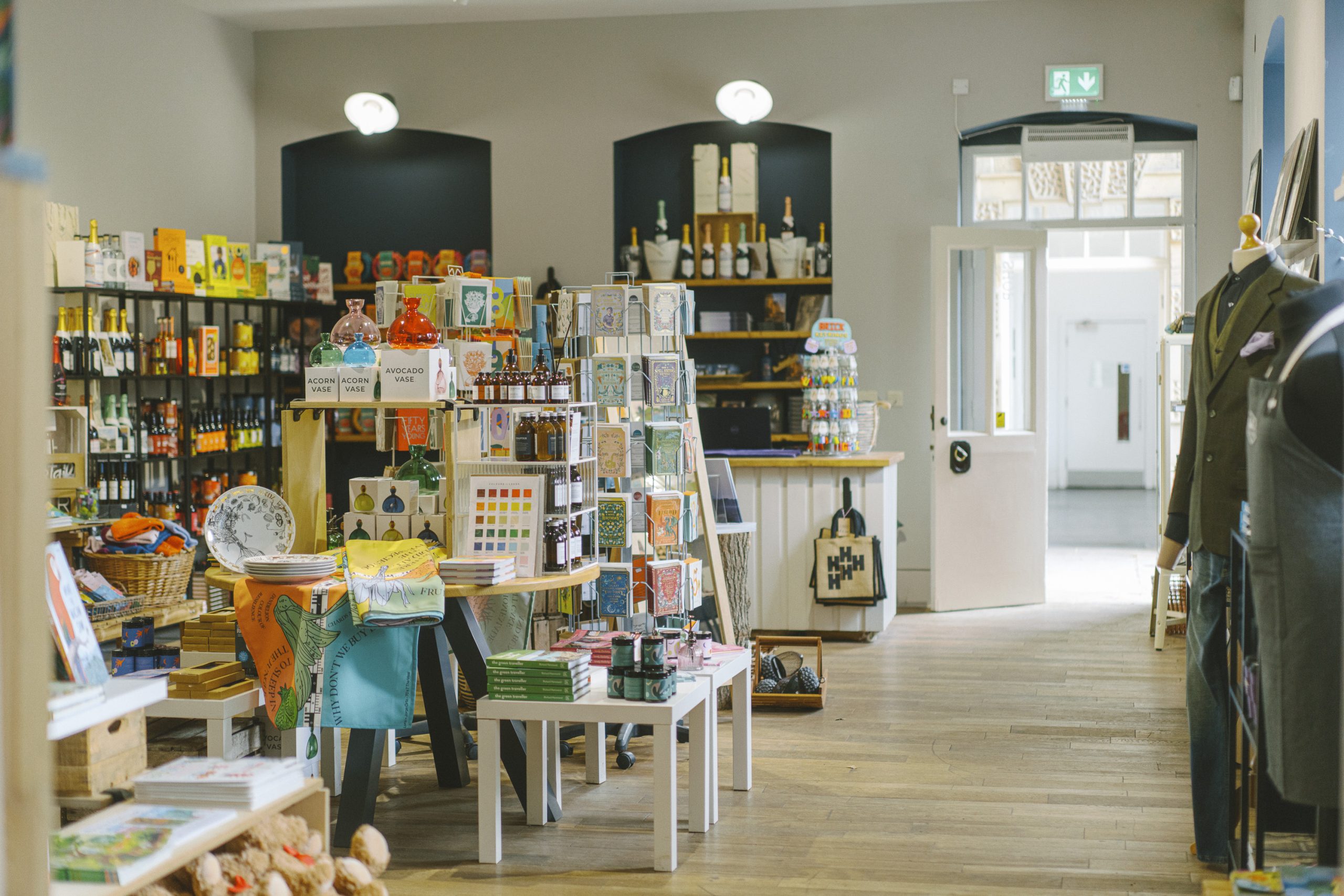 View of the Courtyard Shop from the Second Handbook Shop with official Emmerdale costumes to the left and tables adorned with gift cards, acorn vases, plates, books and bath products. In the background out of focus stands a long shelving unit with bottles of Nyetimber sparkling wine, Harewood Beer and Harewood Gin.